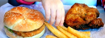 Mano di bambina seduta davanti a hamburger e patatine frette, ricche di acrilammide
