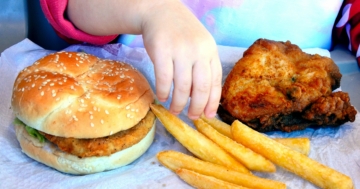 Mano di bambina seduta davanti a hamburger e patatine frette, ricche di acrilammide
