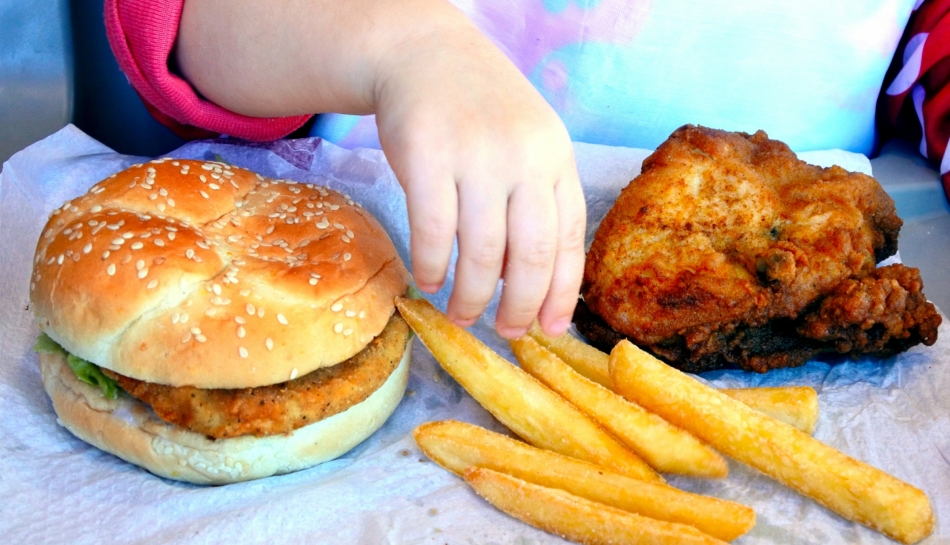 Mano di bambina seduta davanti a hamburger e patatine frette, ricche di acrilammide