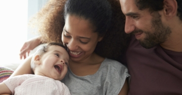 Mamma e papà seduti sul divano abbracciano la loro bambina che ha meno di mille giorni di vita