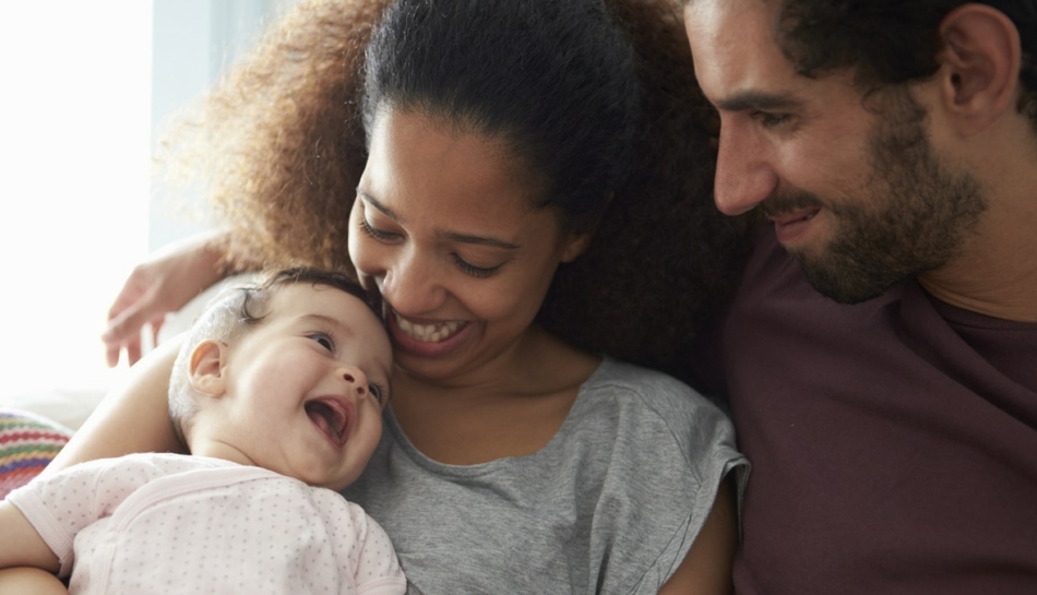 Mamma e papà seduti sul divano abbracciano la loro bambina che ha meno di mille giorni di vita