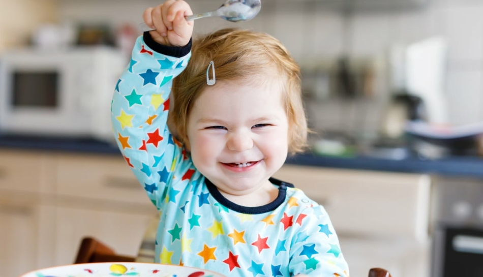 Bambina piccola che agita ridendo il cucchiaio prima di mangiare, in un'atmosfera serena che evita lo sviluppo di disturbi alimentari